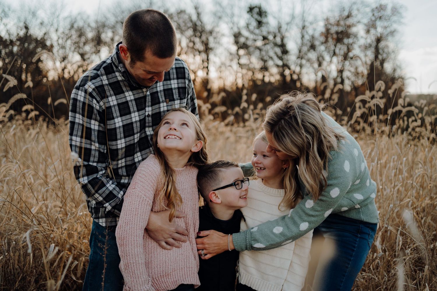 Family hugging their three young children in the tall grass.