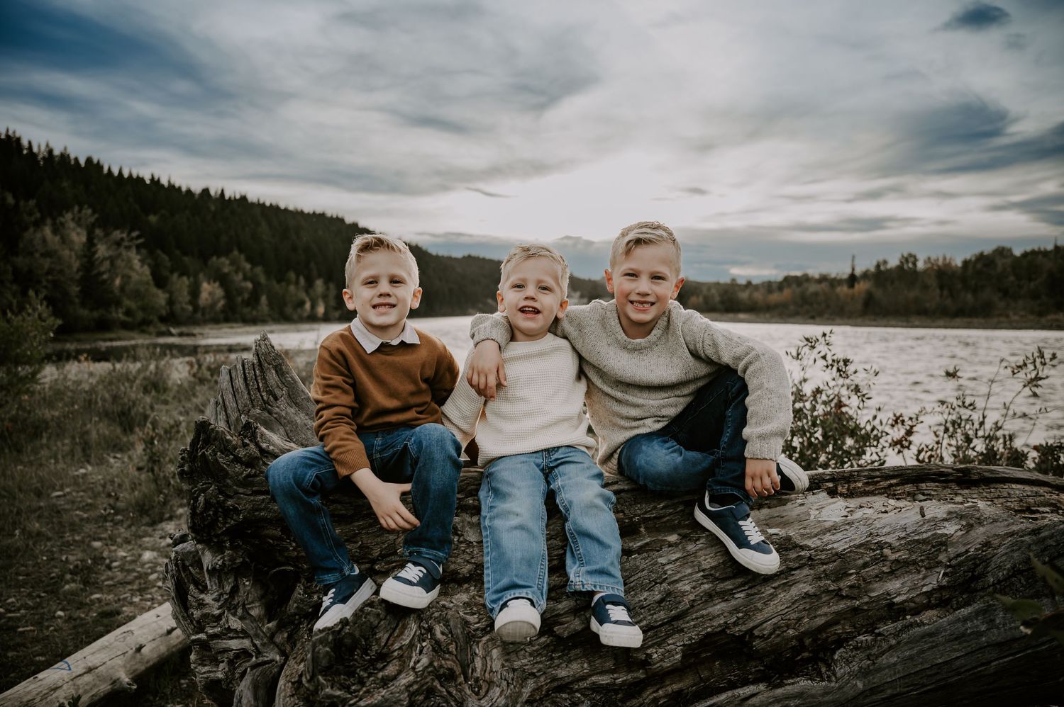 Three young children sit together on a fallen log by a lake with mountains in the background.