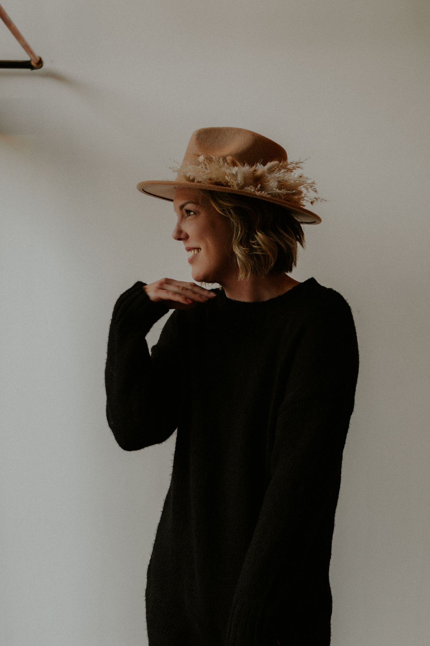Stylish figure in a black sweater wearing a brown felt hat decorated with neutral dried flowers.