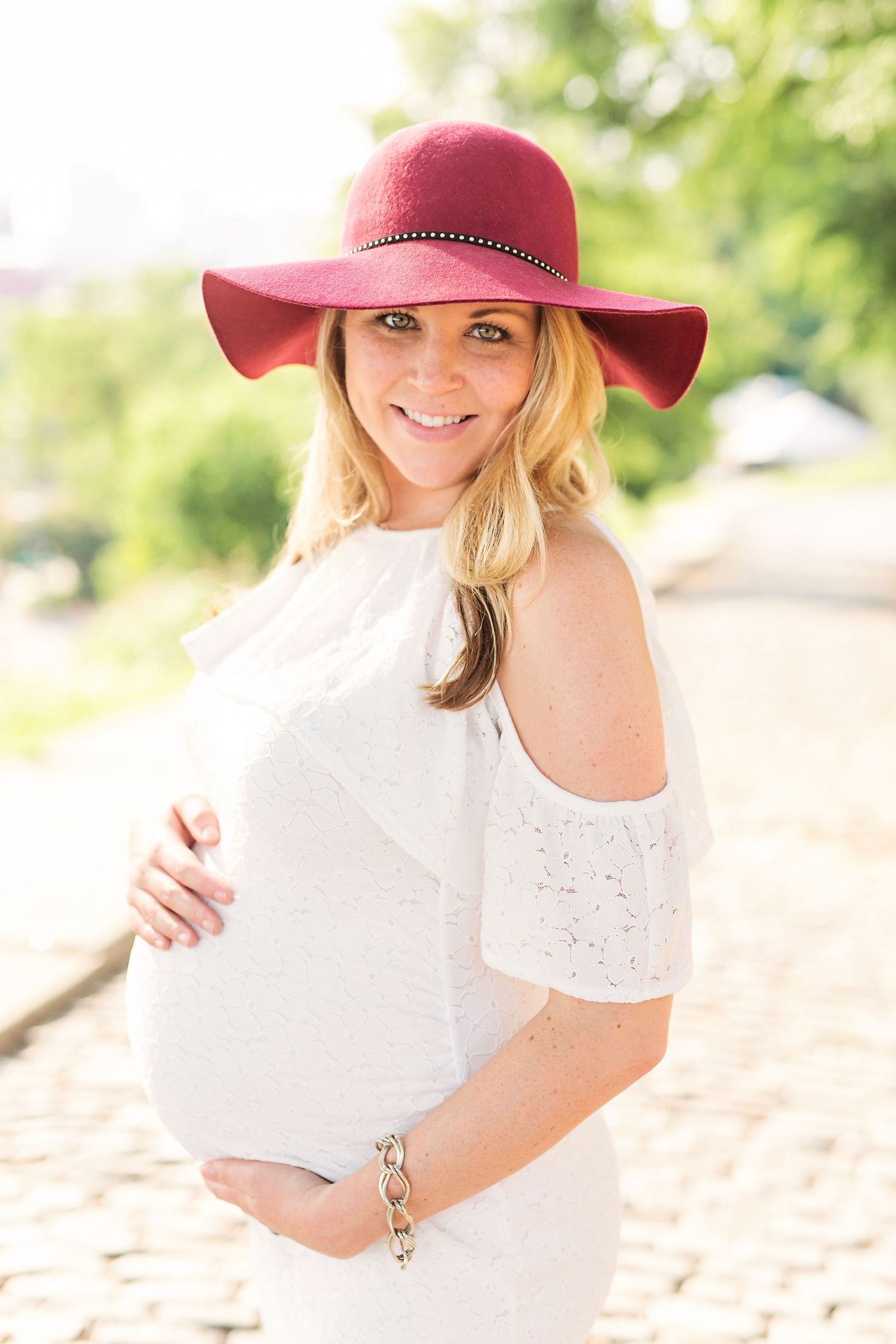 Woman in a burgundy hat and white dress poses outdoors during pregnancy photoshoot.