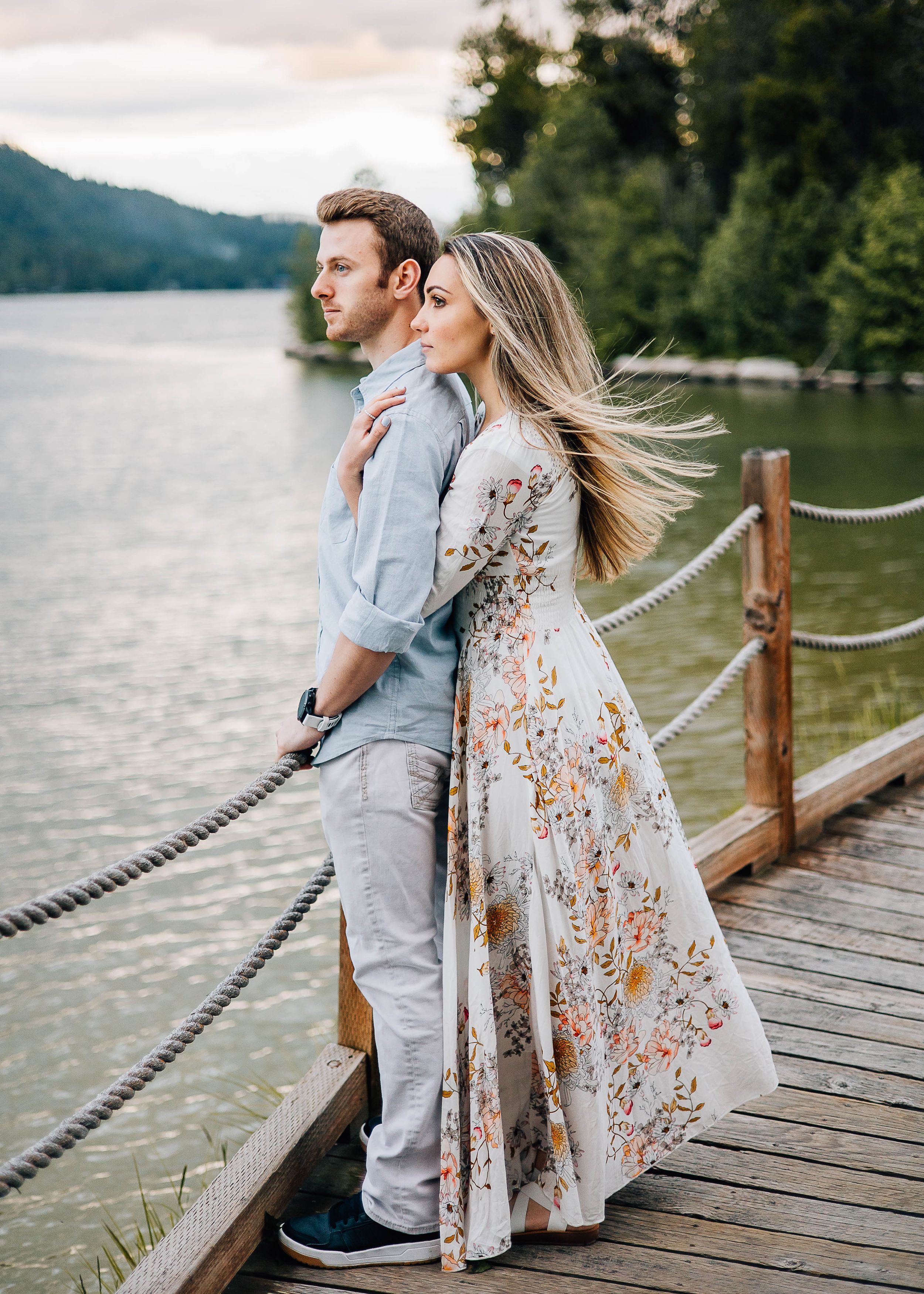 A couple stands on a wooden dock overlooking a mountain lake at dusk.