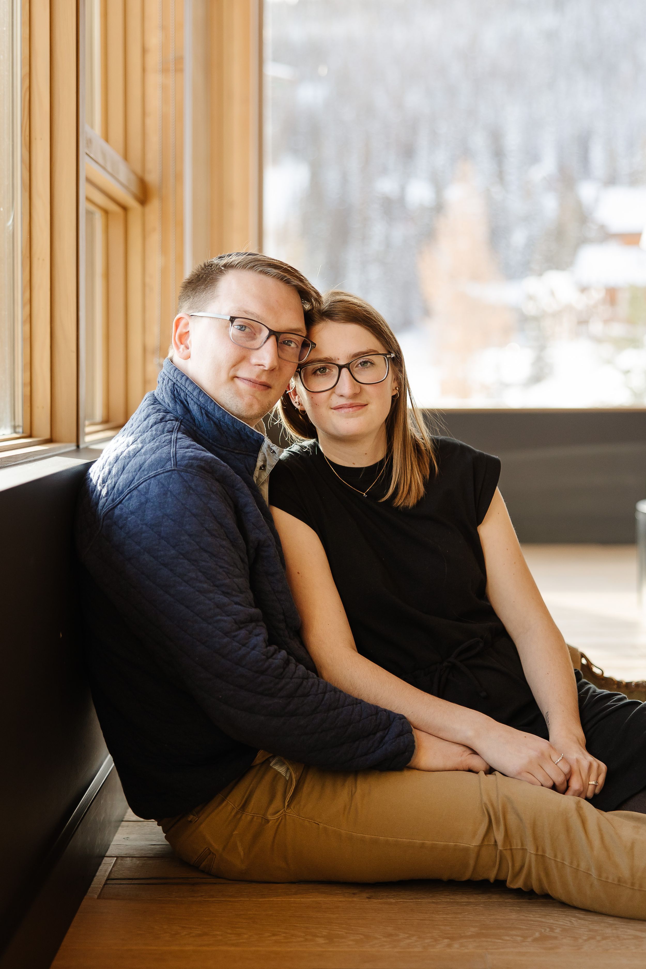 Couple sitting by window in cozy winter scene wearing matching glasses.