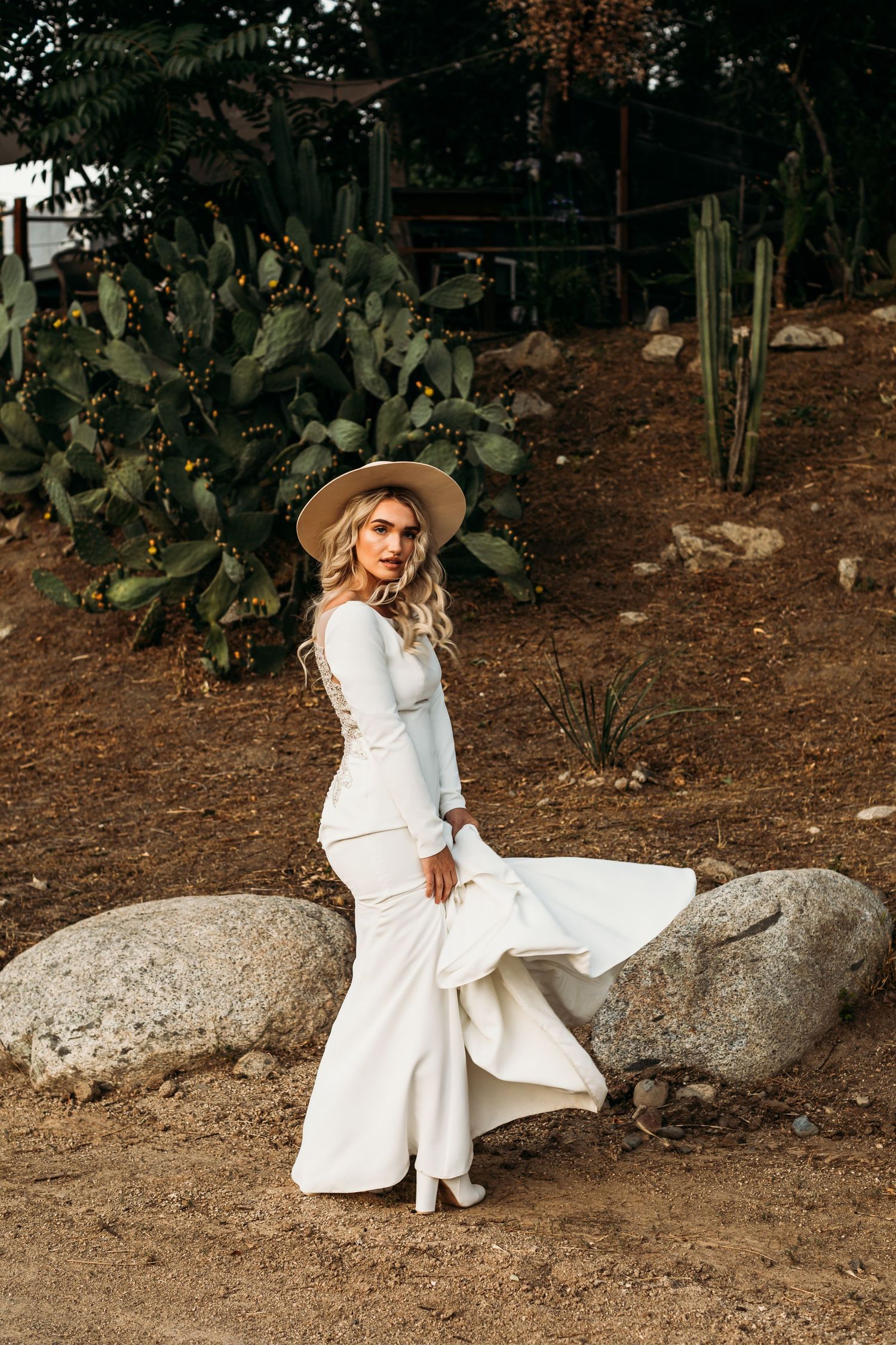 A woman in a white dress and hat poses elegantly among desert cacti and rocks.
