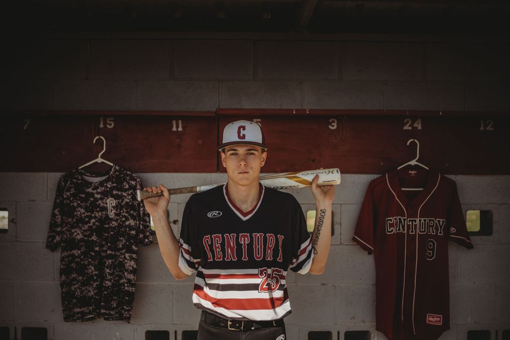 Teen baseball player poses in locker room holding bat across shoulders with jerseys hanging behind.