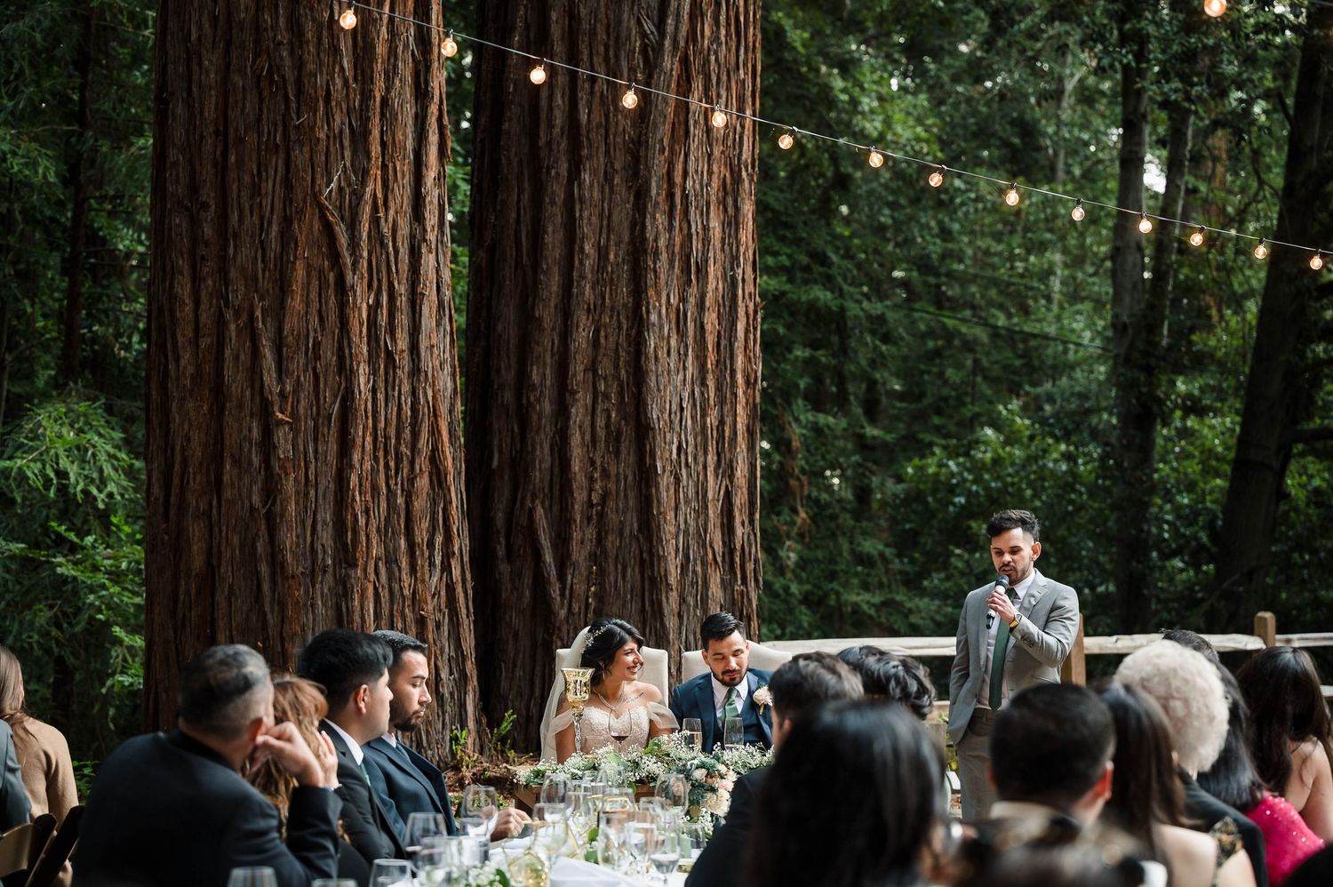 Forest wedding reception set among towering redwood trees with string lights overhead.