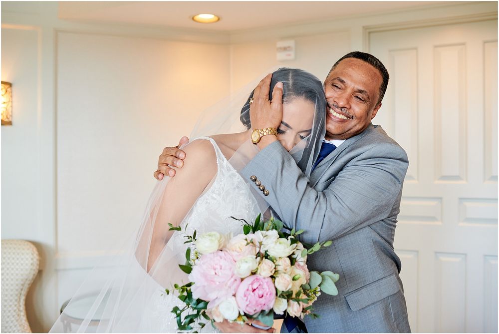 A bride in a white gown and veil stands in an elegant room with cream colored chairs.
