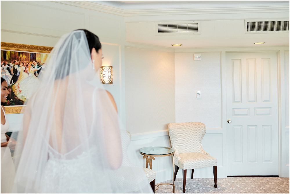 Bridesmaids in light colored dresses celebrate excitedly in a bright hotel room.
