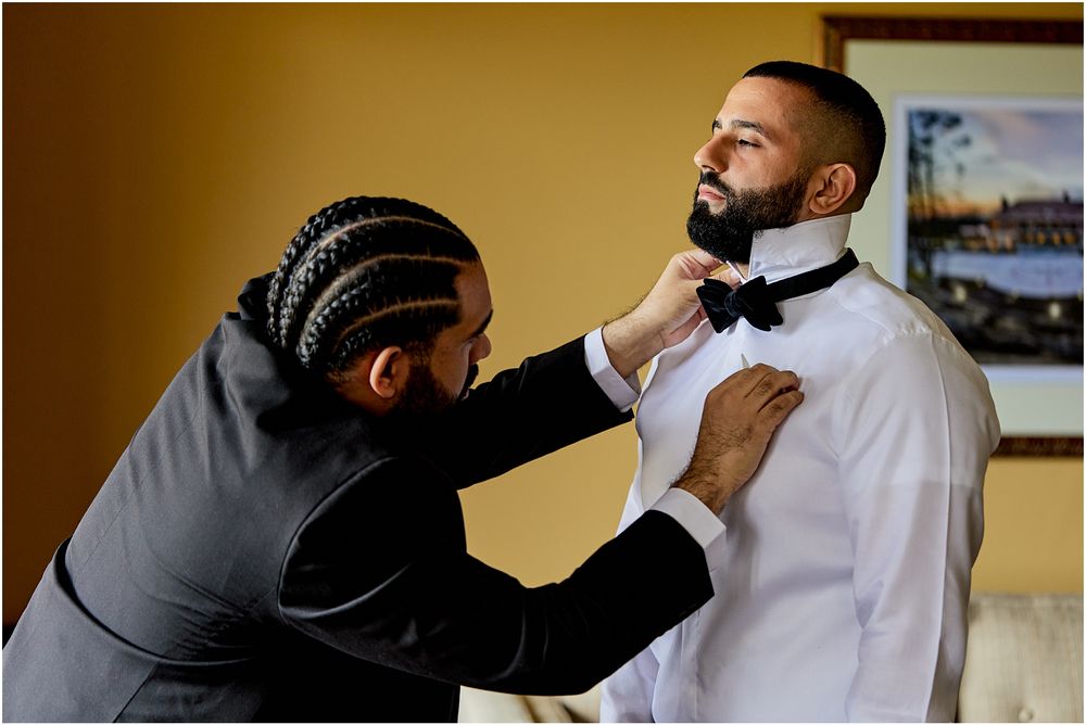 Two people adjusting a formal black bow tie against a warm-toned wall.