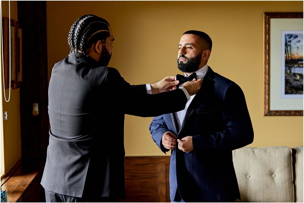 Two people making final adjustments to a navy blue tuxedo and bow tie.