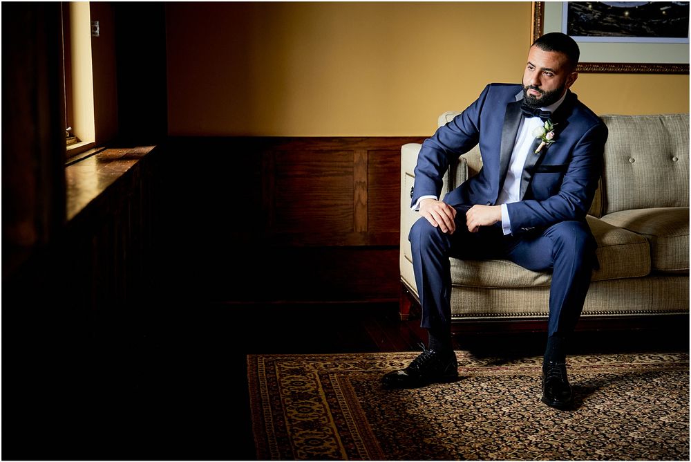Man in navy tuxedo sitting on beige couch near window with dramatic lighting.