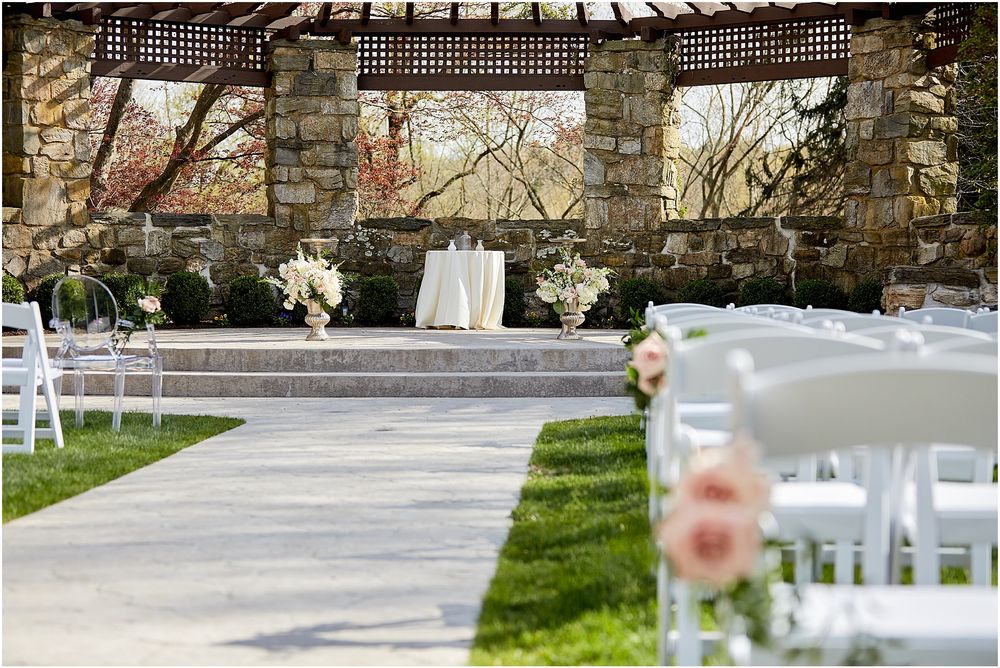 Outdoor wedding ceremony setup with stone archway and white chairs on grass.