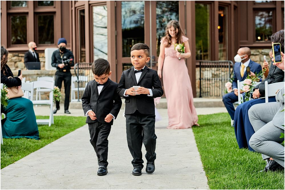 Two young ring bearers in black suits walking down outdoor ceremony aisle.