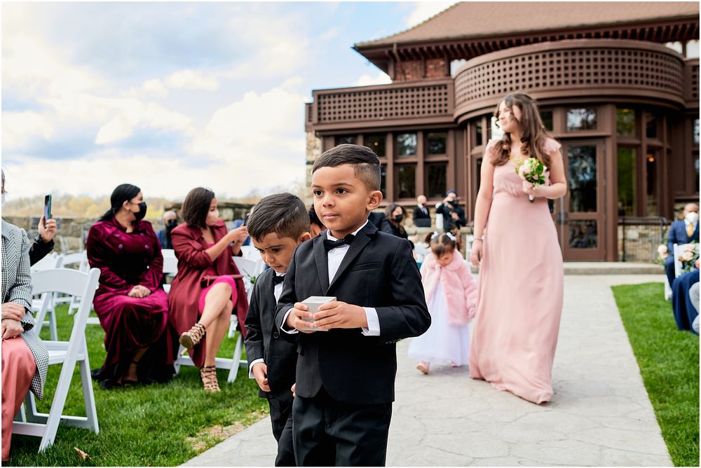 Young ring bearer in tuxedo walks down outdoor wedding aisle as bridesmaid in pink dress follows behind.