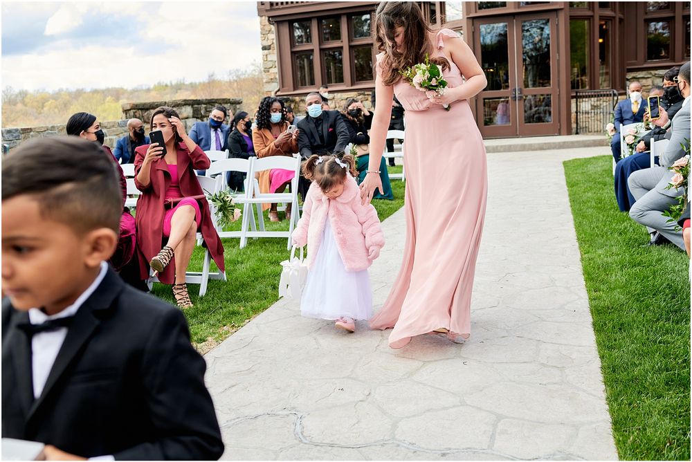 Outdoor wedding ceremony with bridesmaid in pink gown and flower girl walking down stone pathway.