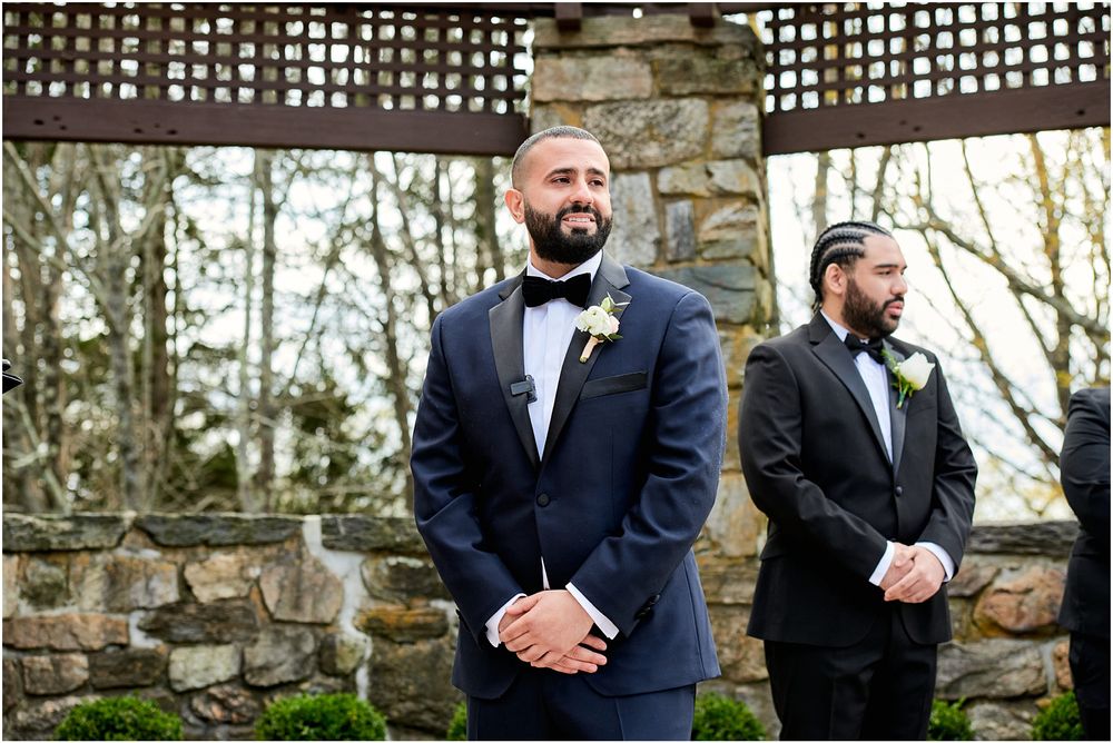 Groomsmen in navy tuxedos stand under stone pergola during outdoor wedding ceremony.