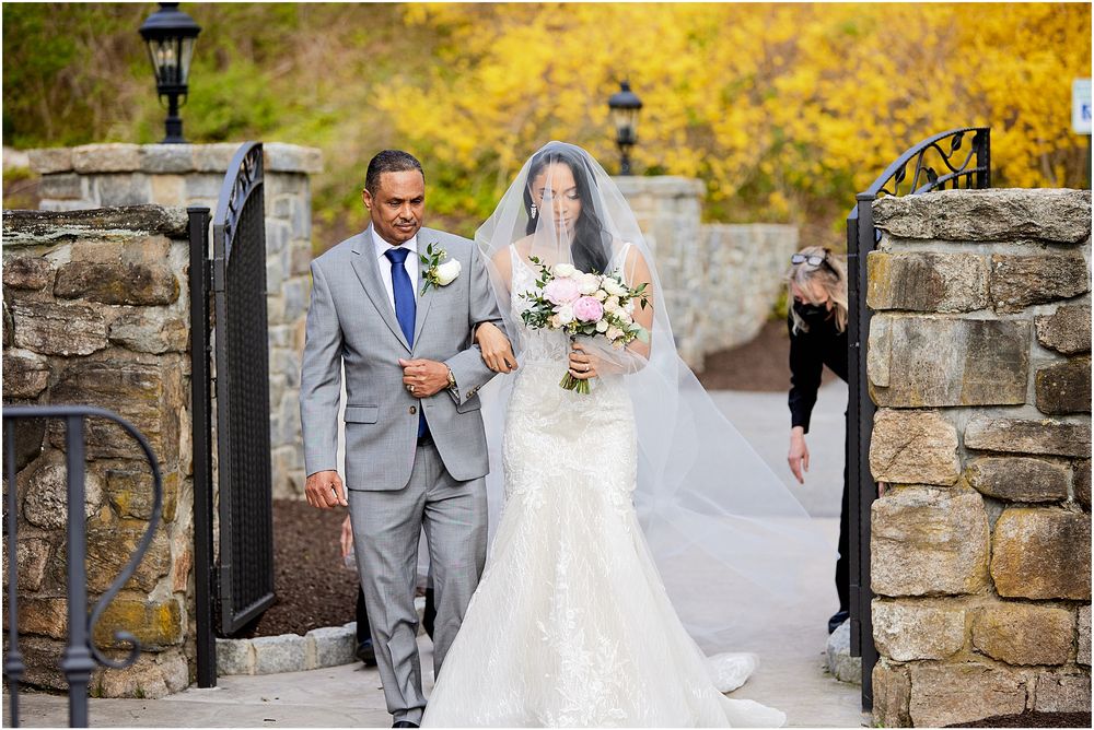 Bride in lace wedding gown walks along stone pathway carrying autumn floral bouquet.