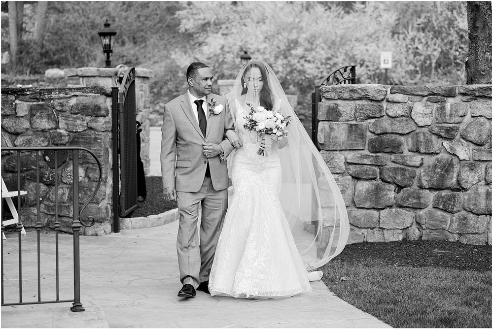 Black and white photo of couple walking along stone pathway during wedding ceremony.