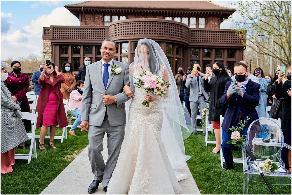 Wedding ceremony processional on stone pathway with guests seated in white chairs.