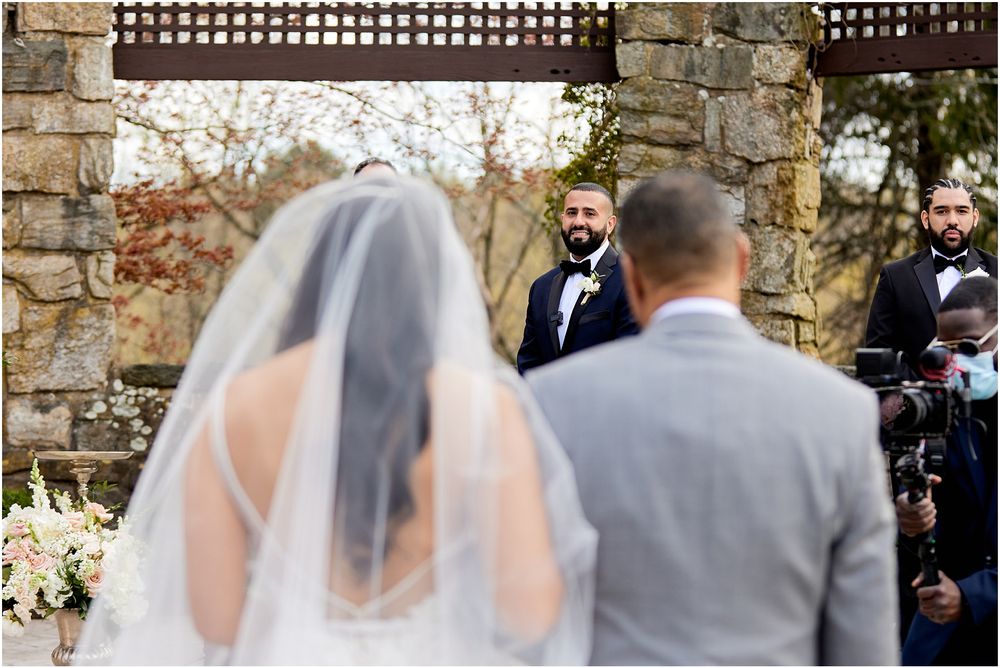 Wedding ceremony at stone venue with guests seated on white chairs outdoors.