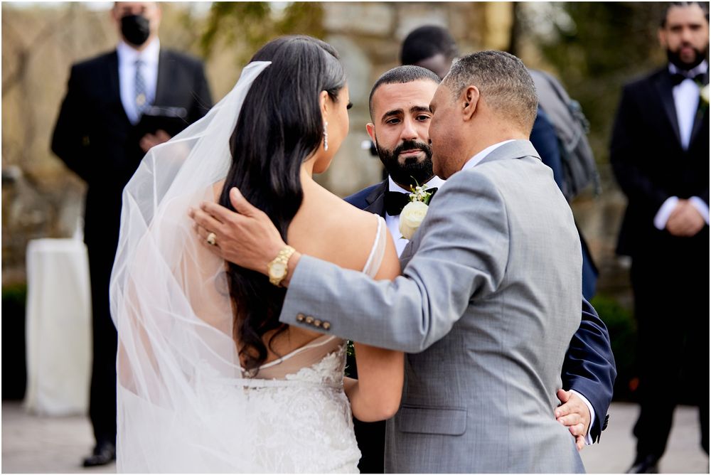 Intimate wedding ceremony moment with stone architecture and floral details in background.