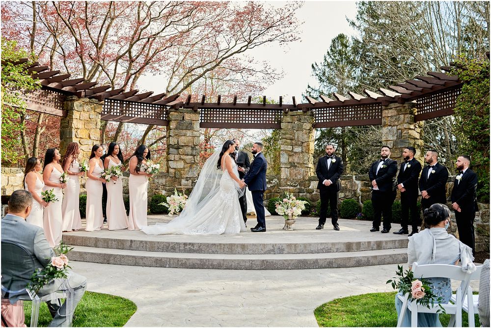 Wedding ceremony under a wooden pergola with stone pillars and spring blossoms in the background.