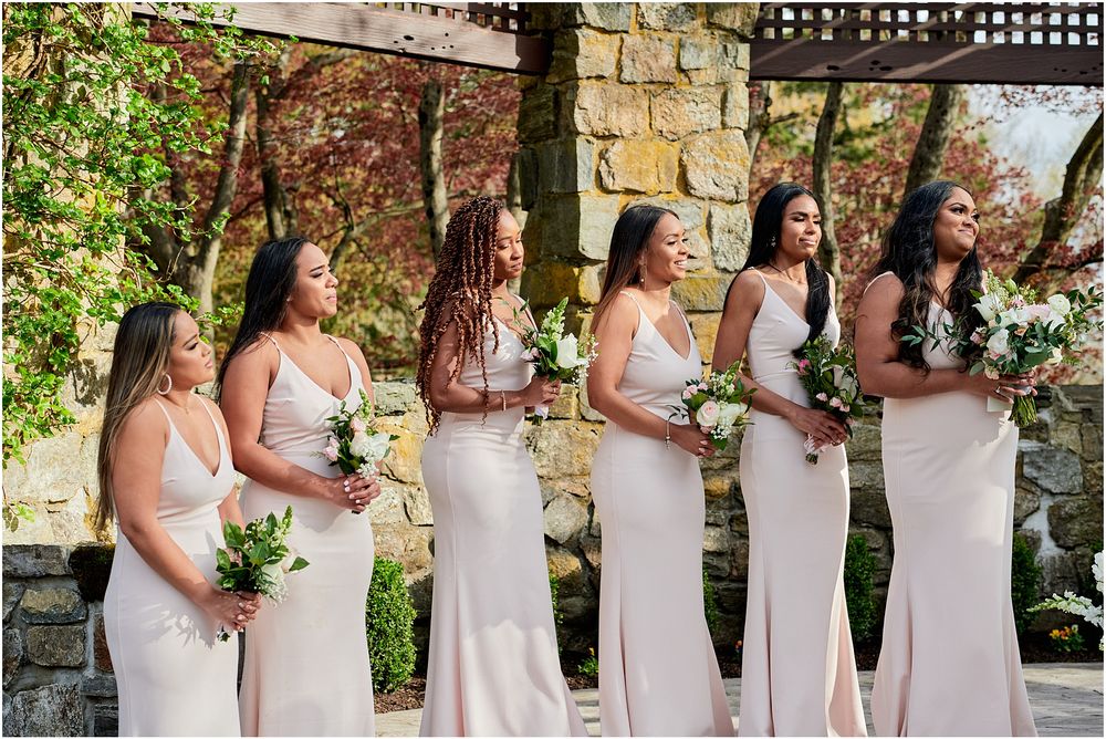 Row of bridesmaids in blush pink dresses holding bouquets against a stone wall backdrop.
