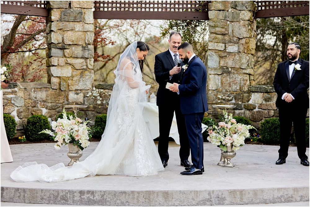 Wedding ceremony in front of a rustic stone wall with floral arrangements and wooden pergola.