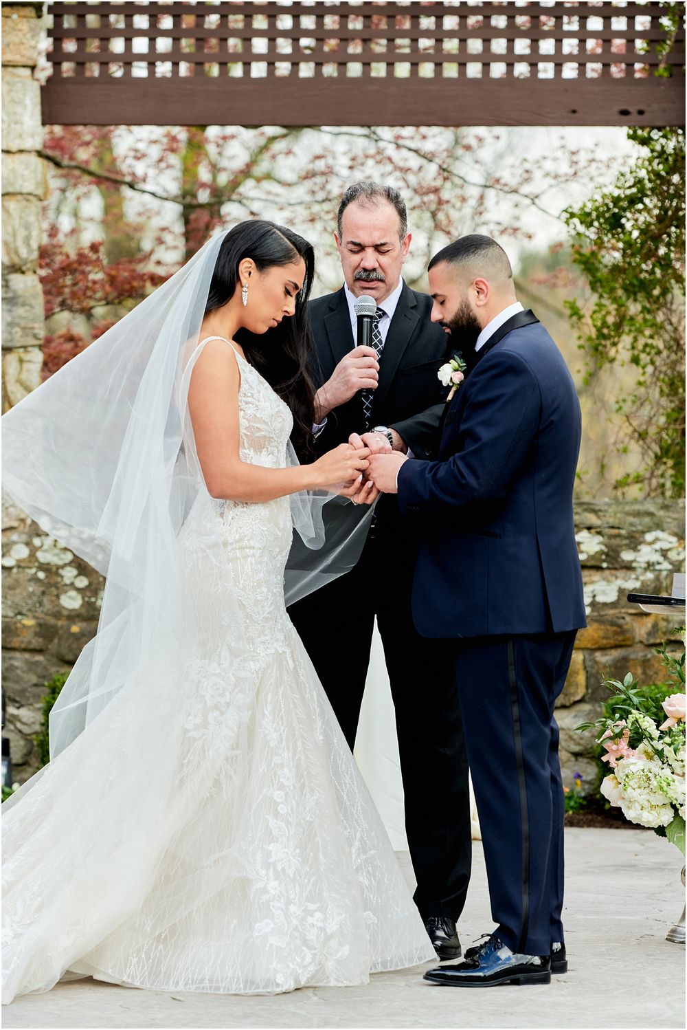 Ring exchange ceremony during outdoor wedding with stone wall and wooden trellis backdrop.