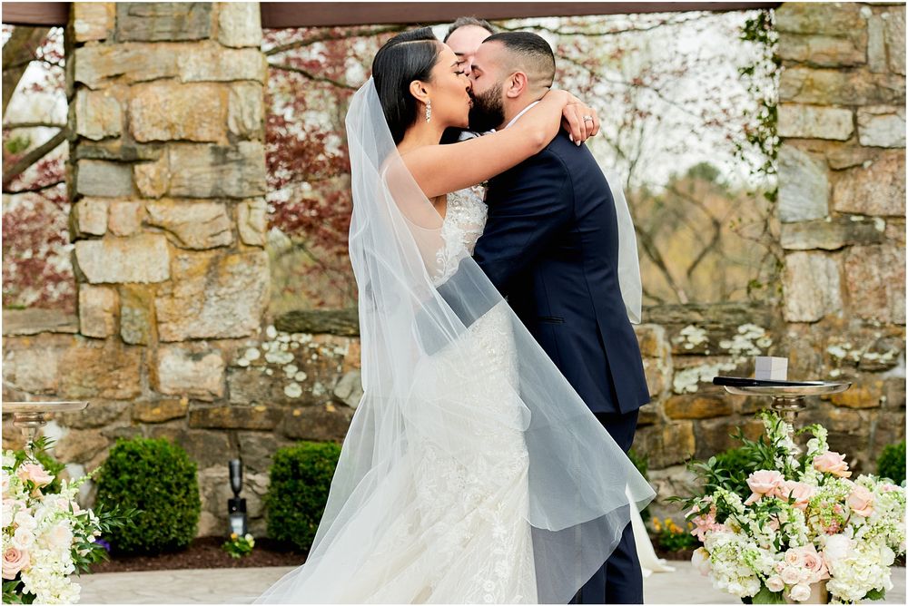 Romantic moment during outdoor wedding ceremony with stone architecture and white floral decor.