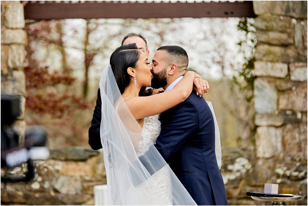 Intimate wedding moment captured against rustic stone wall and wooden pergola.