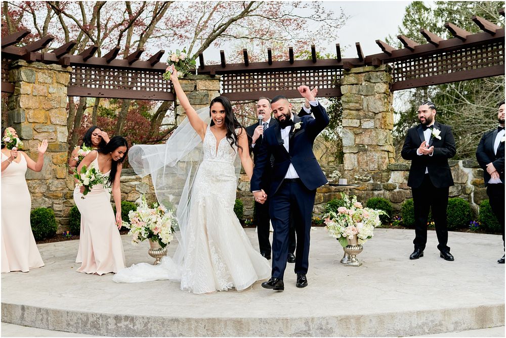 Celebratory moment during outdoor wedding ceremony with stone architecture and spring blooms.