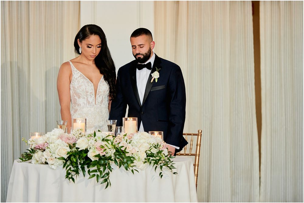 A couple stands at their wedding reception sweetheart table decorated with elegant white floral arrangements.