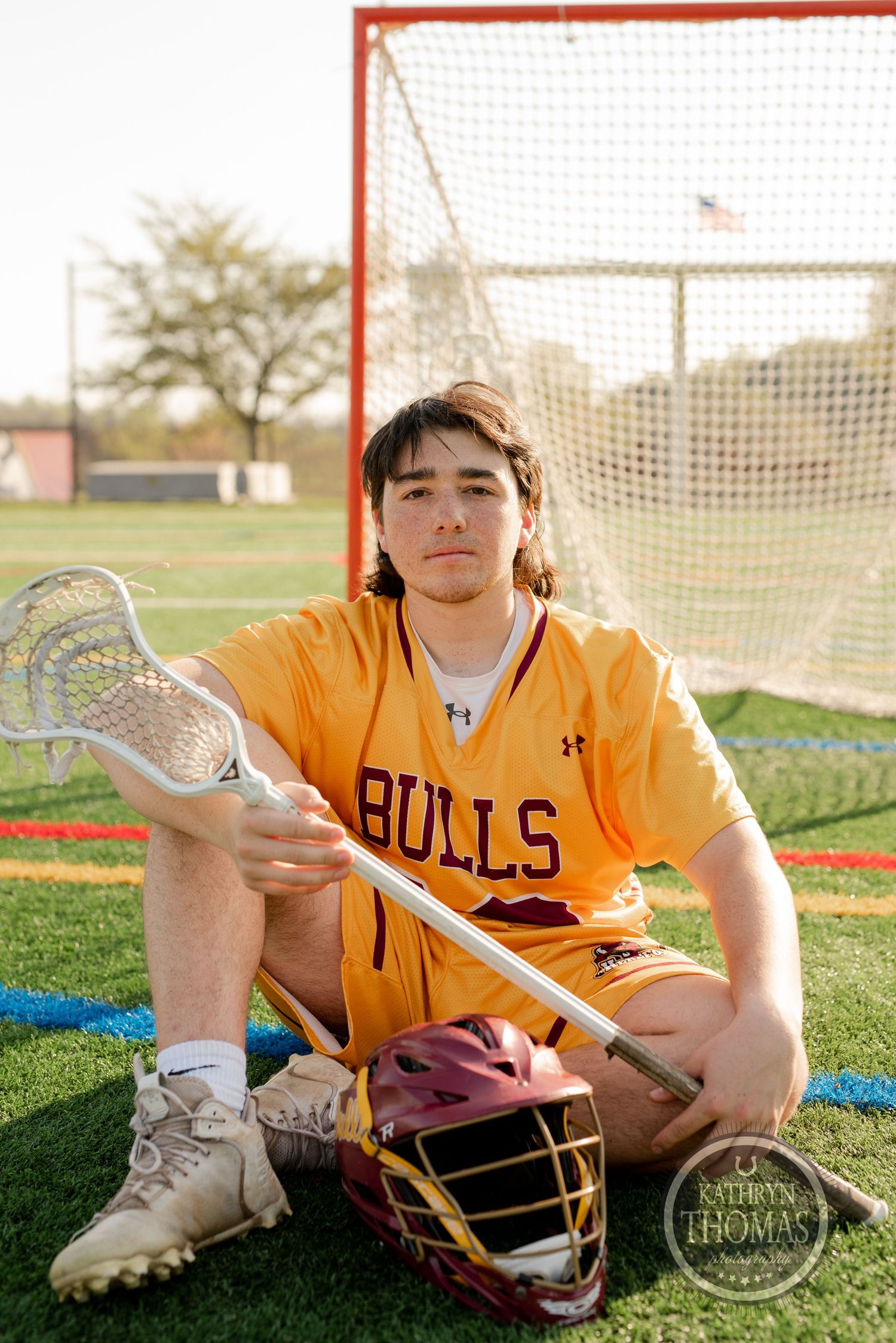 Lacrosse player in Bulls uniform holding stick while sitting by goal net on field.