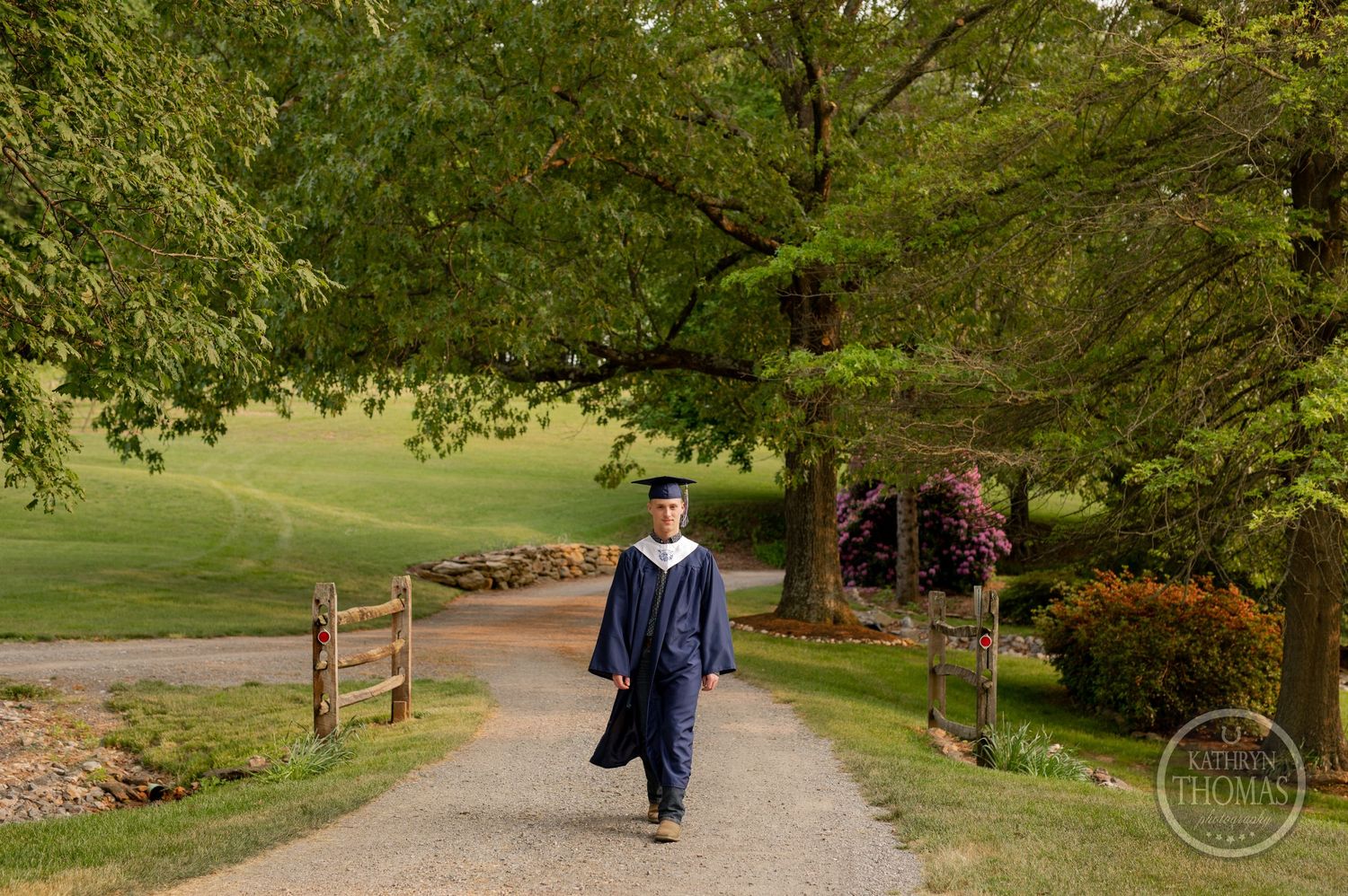 Graduate in academic regalia walks down a scenic country path between wooden fences.