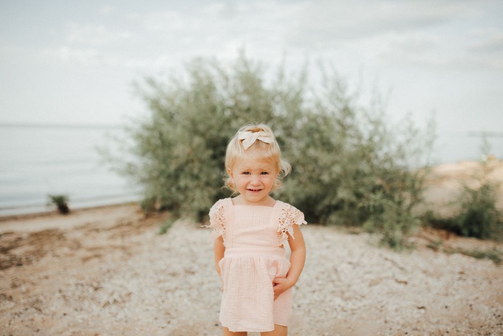A toddler in a pink dress stands on a beach with shrubs and water in the background.