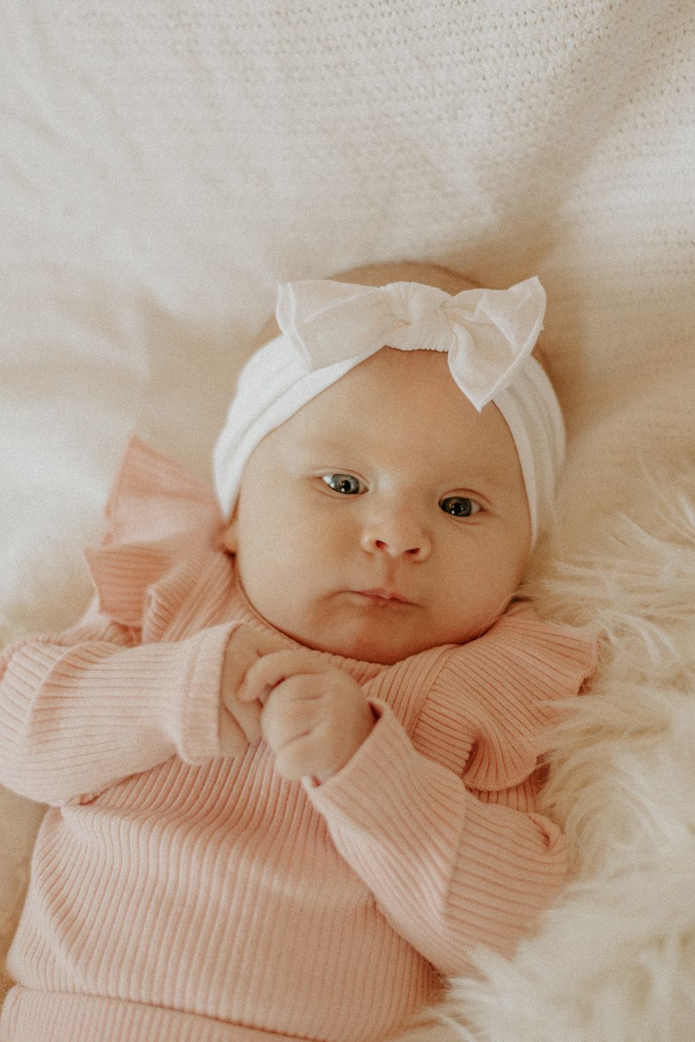 Baby wearing pink ribbed outfit and white bow headband lying on soft white fabric.