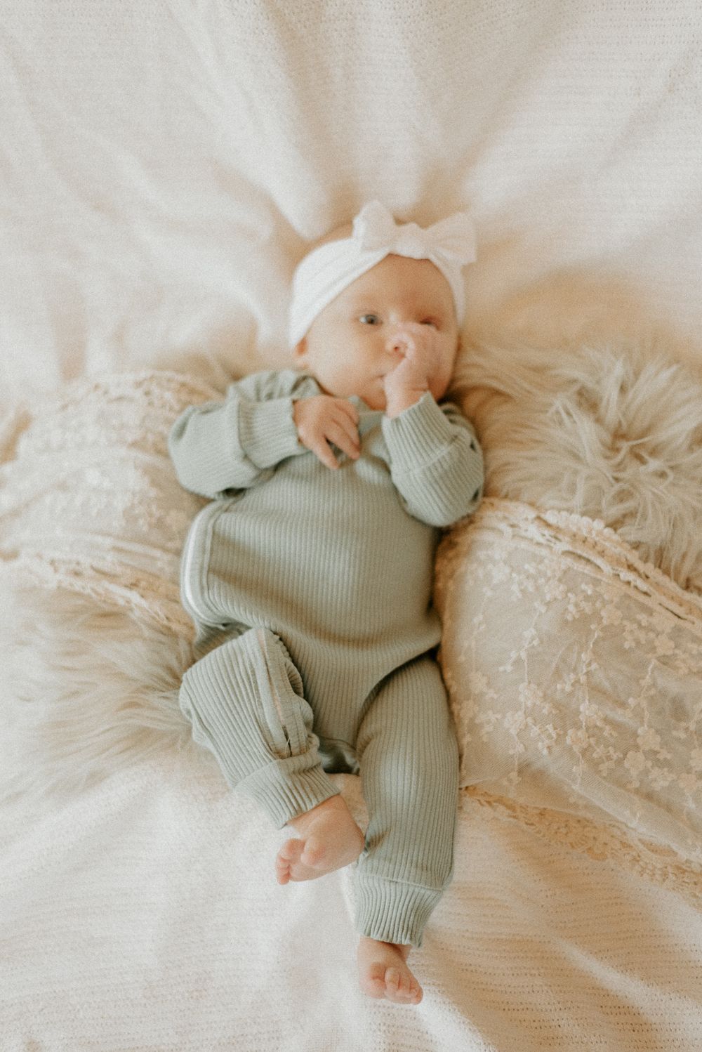 Baby in sage green ribbed outfit with white bow headband resting on white textured bedding.