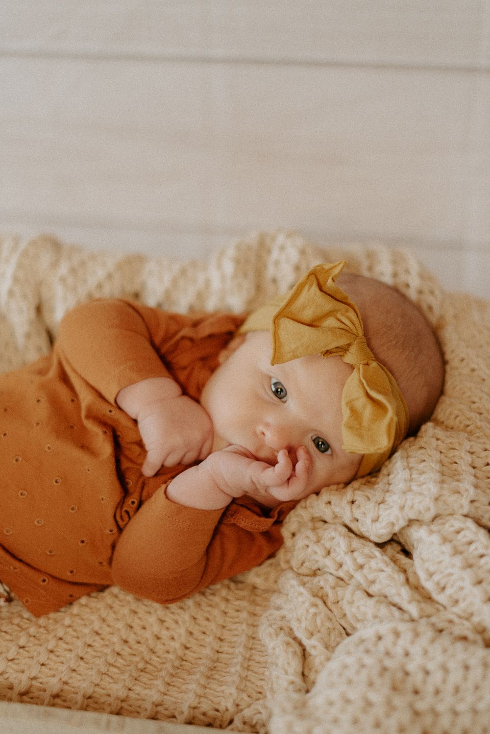 Baby in rust-colored outfit with gold bow headband lying on cream knit blanket.