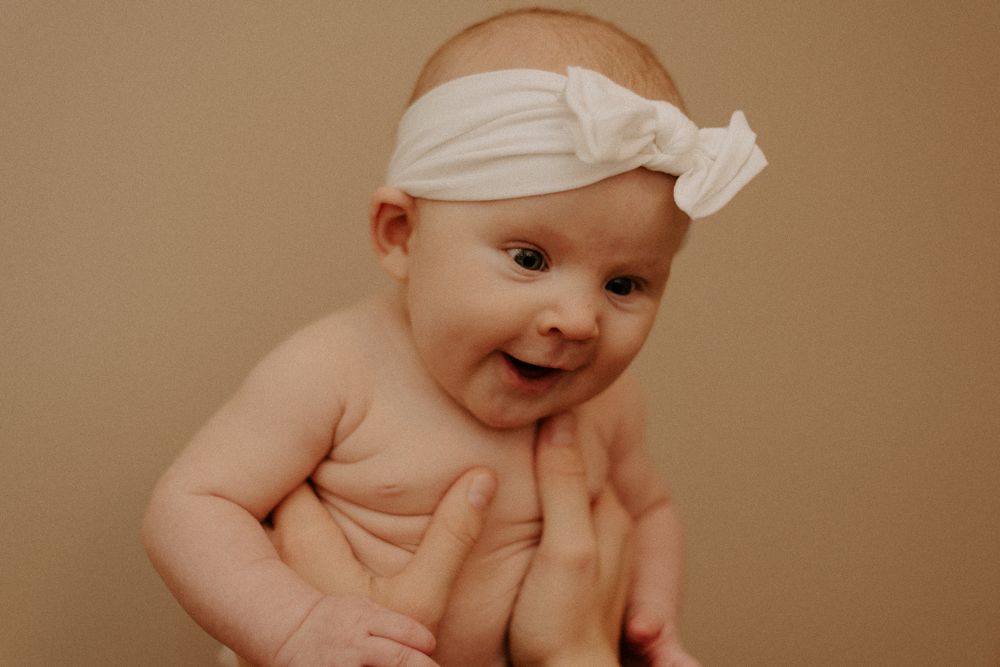 Baby wearing white bow headband smiling on neutral background.
