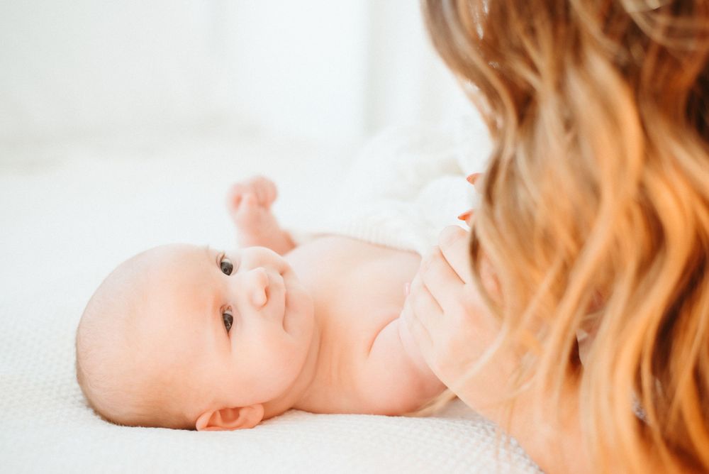 A small infant lying on white bedding reaches towards the camera with curious eyes.