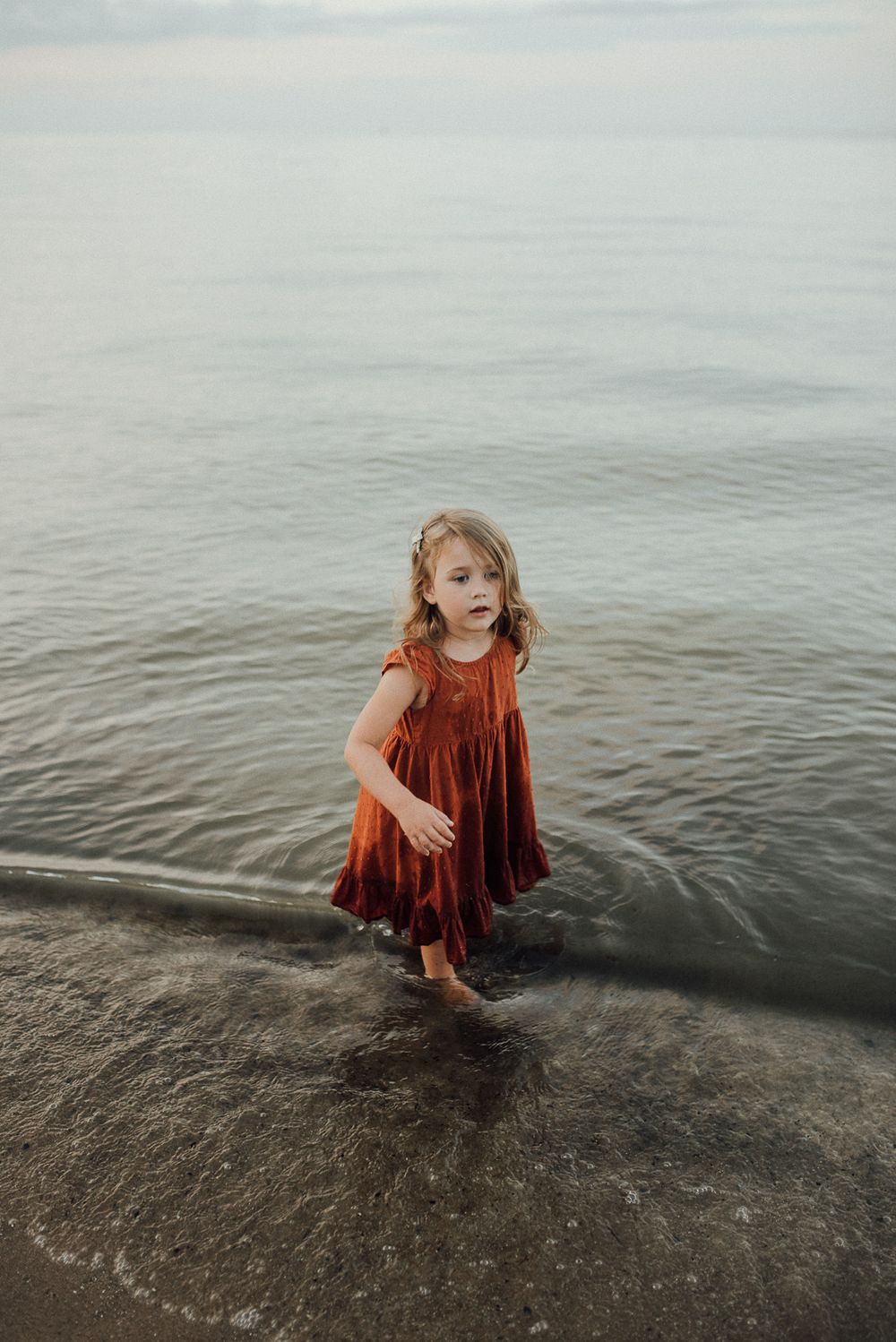 A young child in a red velvet dress plays in the shallow water at the beach.