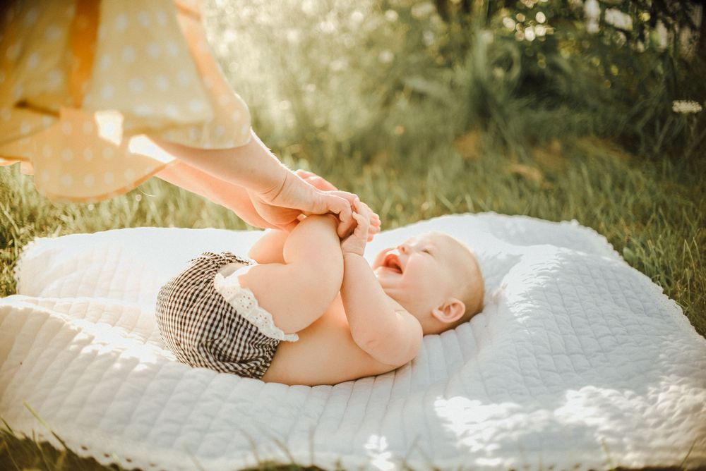 Baby in knit shorts lying on white blanket in sunlit grass while being tickled.