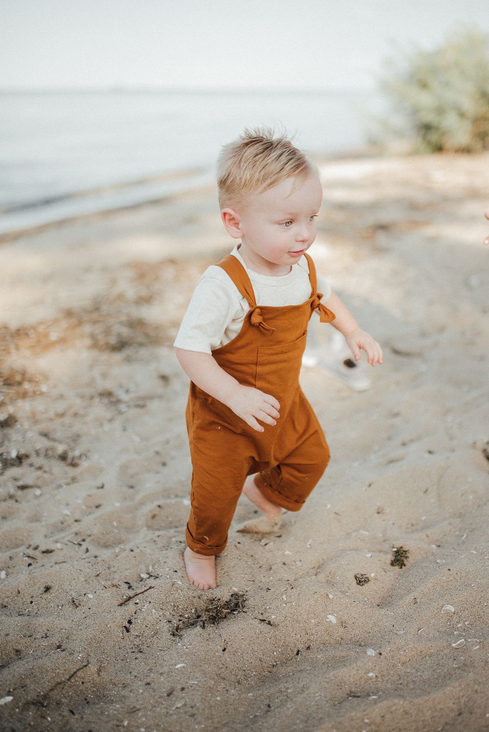 A toddler in brown overalls plays on the sandy beach near the water.