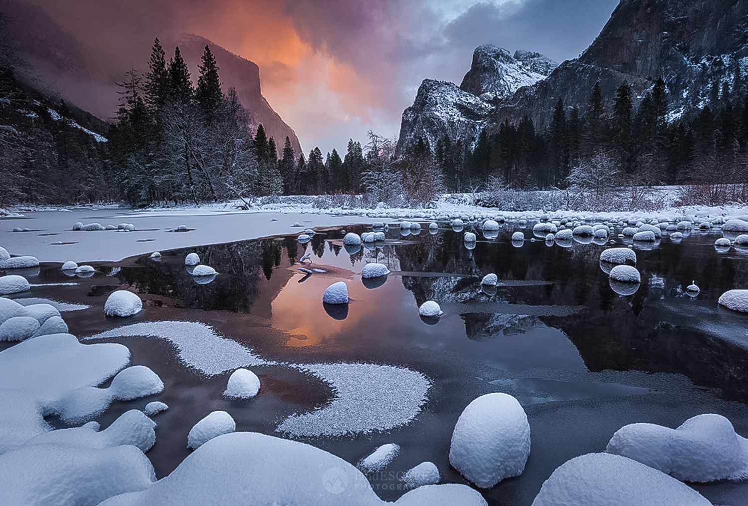 Snow-covered rocks reflect winter sunset at Yosemite Valley with Half Dome and mountain peaks in dramatic twilight sky.
