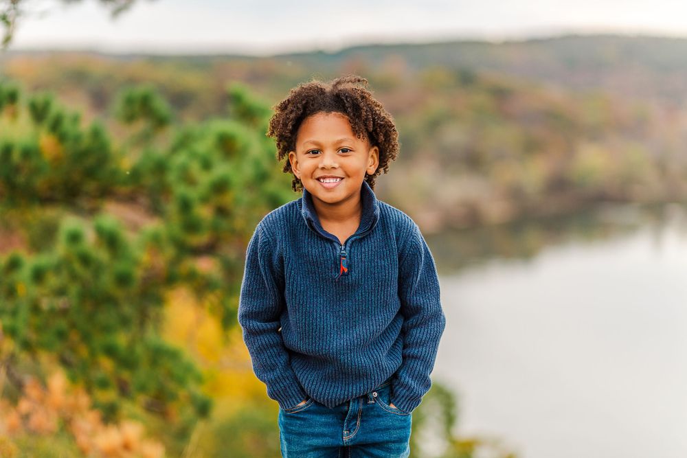 Child wearing navy sweater standing on rocky outcrop with scenic lake view in background.