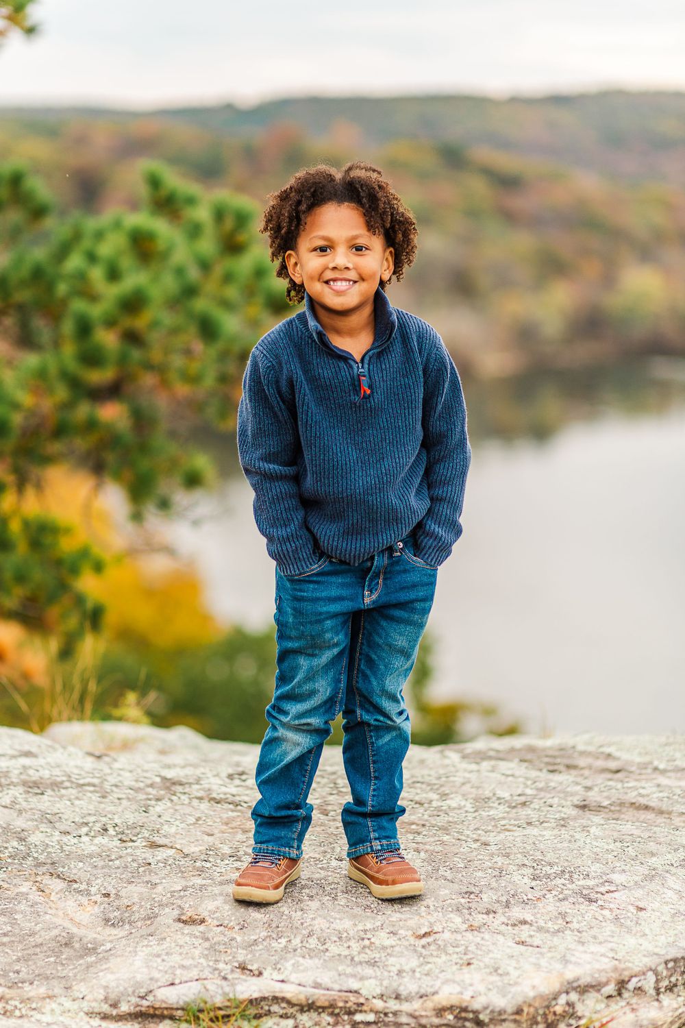 Portrait of young person in blue sweater and jeans standing on rock with autumn scenery behind.