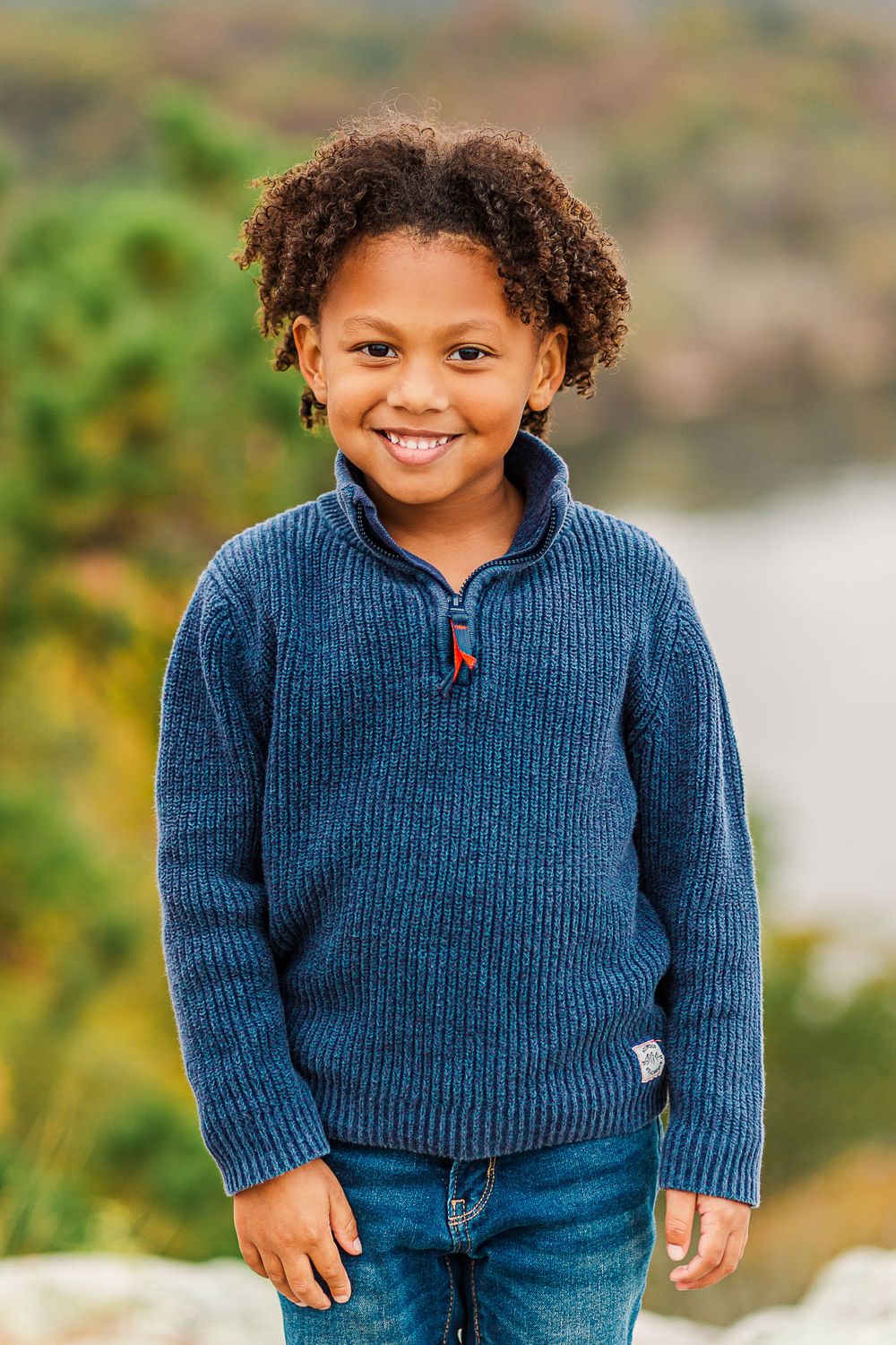 Young person in blue knit sweater and jeans smiling against autumn landscape backdrop.