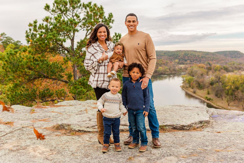 A happy family of four poses together on a rocky overlook with a river and autumn scenery behind them.