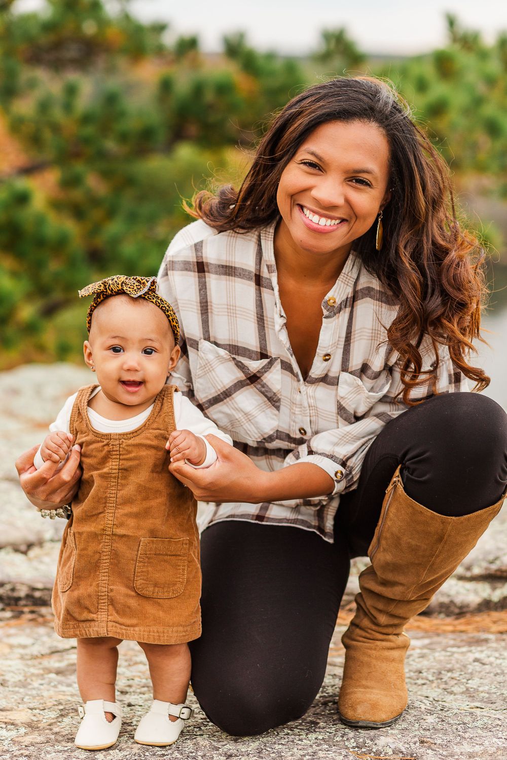 Family portrait with parent in plaid shirt and boots kneeling next to toddler in brown dress on rocky terrain.