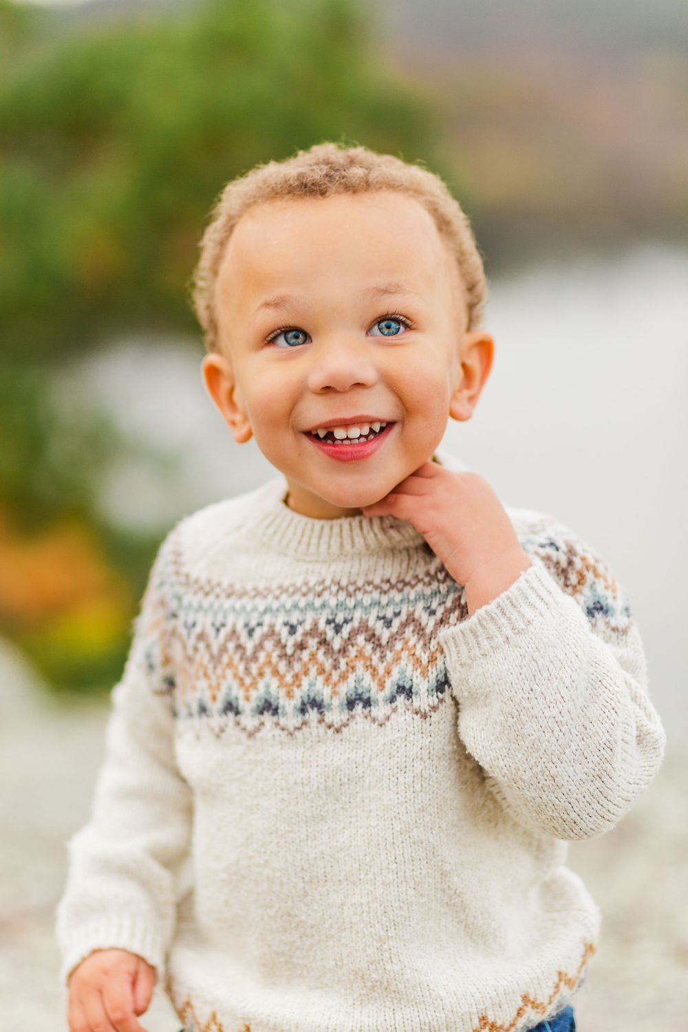 Young child wearing white Fair Isle sweater smiles joyfully at camera in outdoor autumn setting.